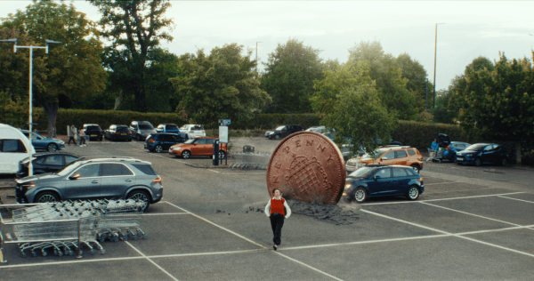 Giant penny rolling through a supermarket car park